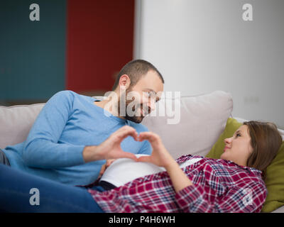 Felice l'uomo e la donna incinta che mostra segni di cuore con le dita mentre vi rilassate sul divano di casa Foto Stock
