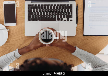 Vista dall'alto di un imprenditore donna seduta con il computer portatile e il telefono cellulare sulla tabella tenendo una tazza di caffè in mano. Imprenditrice di lavoro a casa wi Foto Stock