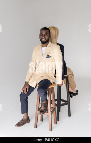 Elegante multietnica giovane coppia seduta di schiena, uomo sorridente in telecamera isolato su grigio Foto Stock