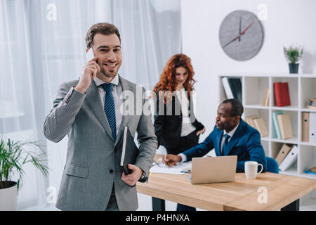 Imprenditore sorridente parlando su smartphone e due colleghi di lavoro lavorare con documenti Foto Stock