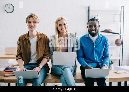 Sorridente imprenditori multiculturale utilizzando computer portatili e seduta sul tavolo in ufficio moderno Foto Stock