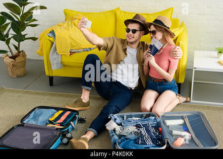 Bella giovane tenendo selfie sullo smartphone mentre valigie di imballaggio per le vacanze Foto Stock