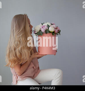 Felice donna bionda con fiori in un cappello box. Bouquet di peonie. Ragazza in un morbido abito rosa con una rosa bouquet di peonie Foto Stock