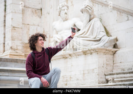 Bel giovane con capelli ricci in tuta tenendo selfie con il suo telefono cellulare nella parte anteriore del dio Nilo statua in Roma Foto Stock