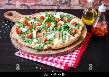 La pizza fatta con una base di pasta acida fresca rucola, pomodorini e formaggio parmigiano su una tavola di legno Foto Stock