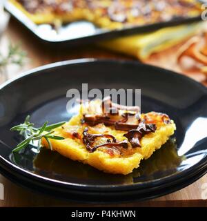 Una fetta di torta di polenta con finferli e cipolle su una piastra di fronte a un vassoio da forno Foto Stock