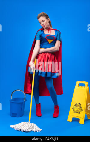 Donna attraente in costume da supereroe con il simbolo di attenzione, benna e mop per pulizia isolato su blu Foto Stock