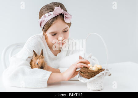 Carino bambina abbracciando il coniglio e guardando il cesto con uova di pasqua nel nido Foto Stock