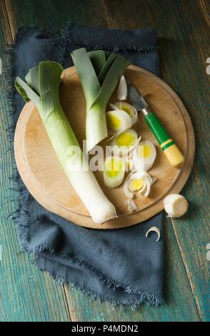 Tettuccio di colpo di due porri su un round tagliere di legno tagliato da un retro in legno coltello gestite e panno blu scuro e verde sullo sfondo di legno Foto Stock