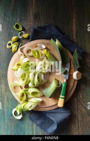 Tettuccio di colpo di porri essendo tritato e preped su un round tagliere di legno da una verde e giallo gestite coltello su un panno blu scuro e verde superficie in legno Foto Stock