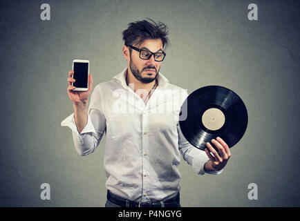 Giovane uomo barbuto in bicchieri guardando perplesso mentre scegliendo tra il disco di vinile e lo smartphone Foto Stock
