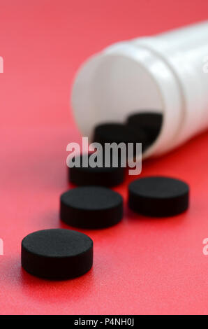 Diverse compresse nero ricade al di fuori del vaso in plastica sulla superficie rossa. Immagine di sfondo sul farmaceutico e medico argomenti. Il carbone attivato . Foto Stock