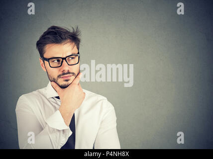 Giovane uomo barbuto in contemplazione guardando verso l'alto e la pianificazione di nuove strategie di business e la creazione di idea su sfondo grigio Foto Stock