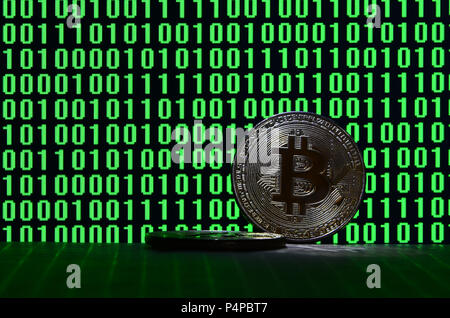 Una coppia di bitcoins giace su di una superficie di cartone sullo sfondo di un monitor raffigurante un codice binario di colore verde brillante di zeri e una unità di un nero b Foto Stock