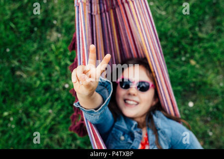 Ragazza sorridente indossando occhiali da sole giacente in amaca mostra segno di vittoria Foto Stock