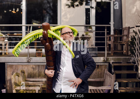 Imprenditore Senior in piedi nel suo giardino, tenendo una gomma palm, sognando di vacanze Foto Stock
