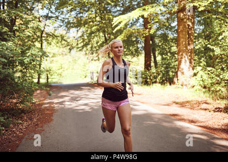 Montare la giovane donna bionda in pantaloncini corti e un tanktop in esecuzione da solo lungo un percorso attraverso una foresta Foto Stock