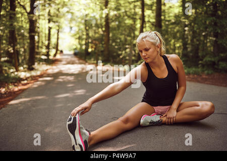 Montare la giovane donna bionda in sportswear seduto sulla strada forestale in fase di riscaldamento con tratti prima di una corsa Foto Stock