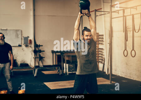 Montare il giovane uomo sorridente sportswear durante l'oscillazione un manubrio sopra la sua testa durante una sessione di allenamento in palestra Foto Stock