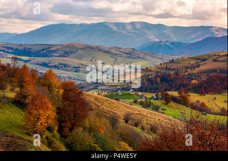 Piccolo villaggio sulle colline in autunno. meraviglioso paesaggio delle montagne dei Carpazi. mighty ridge in distanza. rosso fogliame degli alberi Foto Stock