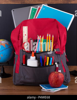 Zaino con un vasto assortimento di forniture scolastiche su una scrivania in legno con un background di Blackboard Foto Stock