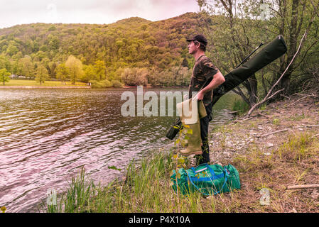 Le avventure di pesca, la pesca alla carpa. Pescatore sulla riva del lago con il camuffamento di attrezzi da pesca, stivali di gomma, sacchetto verde e mimetico multiuso asta Foto Stock