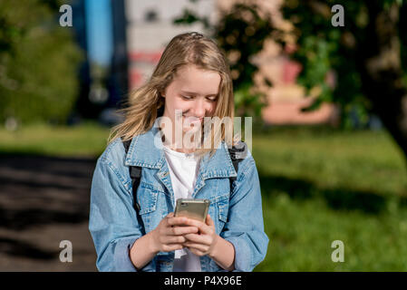 Bambina è bionda. Schoolgirl in estate dopo la scuola. Nelle sue mani detiene uno smartphone. Sorride felicemente. Scrive i messaggi sulle reti sociali. Gioioso riposo dopo l'istituto. Foto Stock