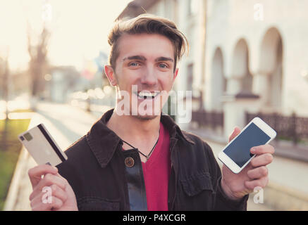 Allegro casual uomo eccitato con la nuova carta fedeltà mentre si tiene lo smartphone e sorridente alla fotocamera su strada Foto Stock