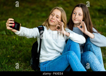 Due ragazze adolescenti. In estate nel parco della natura. Sedersi sul prato dietro gli zaini. Nelle sue mani detiene smartphone. Scattare le foto sul telefono. Il gesto delle mani mostra bene, OK. Il concetto di riconoscimento positivo. Foto Stock