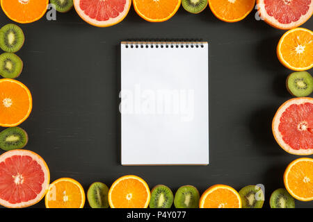 Lay piatto. Vista dall'alto. Notebook con fette di colorata frutta fresca: kiwi, arancia, pompelmo e mandarino su sfondo nero. Estate sfondo. Foto Stock