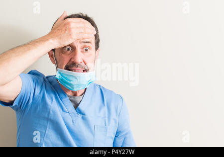 Senior medico uomo utilizzando la maschera ha sottolineato con la mano sulla testa, sconvolto con vergogna e la sorpresa di fronte, arrabbiati e frustrati. Paura e sconvolti per l'errore. Foto Stock