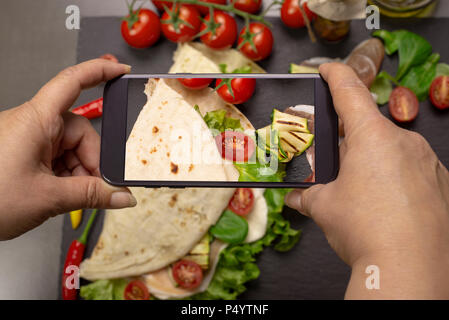 Scattare una foto del cibo con lo smartphone per instagram. Italian piadina romagnola flatbread con lattuga, pomodori ciliegini, il prosciutto crudo, mozzarel Foto Stock