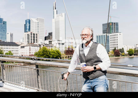 Uomo maturo con la bicicletta e il caffè da asporto sul ponte della città Foto Stock