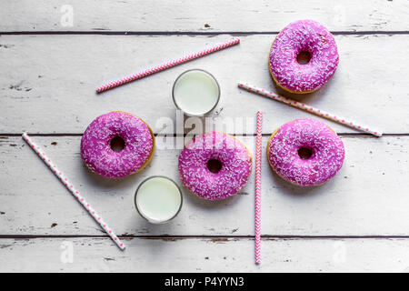 Quattro ciambelle rosa, due bicchieri di latte e le cannucce in legno bianco Foto Stock