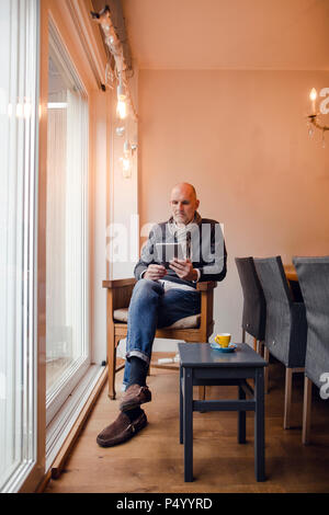 Senior uomo seduto a casa, la lettura sulla tavoletta digitale Foto Stock