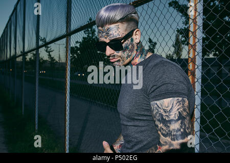Tatuato giovane alla maglia di filo recinzione indossando occhiali da sole Foto Stock