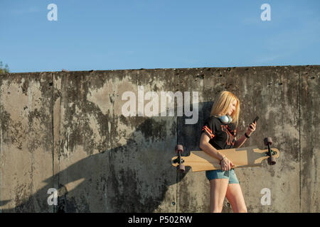 Donna bionda con longboard in piedi nella parte anteriore della parete guardando smartphone Foto Stock