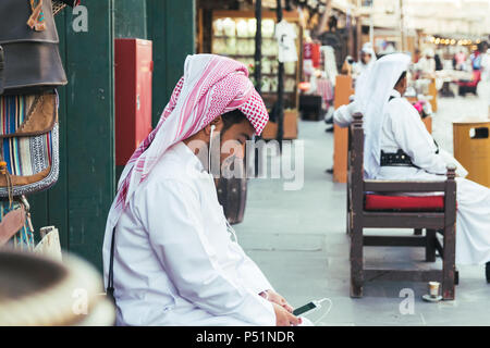 Doha, Qatar - Mart 2018: Arab giovane con keffiyeh sulla sua testa ascoltando la musica sul suo telefono cellulare in corrispondenza della strada. Souq Waqif street a Doha. Foto Stock