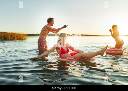 Ridere un gruppo di giovani amici in costume da bagno spruzzi ogni altra divertendosi insieme a un lago al tramonto Foto Stock