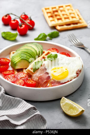 Una sana prima colazione. Cialde belghe con avocado, pancetta e uova fritte in una piastra bianca su un cemento grigio Sfondo. Foto Stock