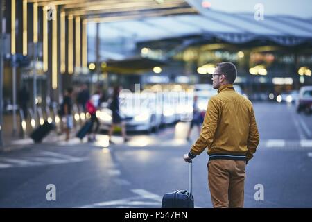 A piedi passeggero al di fuori del terminal dell'aeroporto. Vista posteriore del giovane uomo con i bagagli di notte. Foto Stock