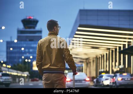 A piedi passeggero al di fuori del terminal dell'aeroporto. Vista posteriore del giovane uomo con i bagagli di notte. Foto Stock