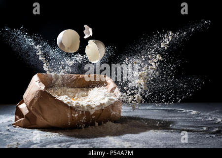 Sacchetto con farina e uova battenti in farina su sfondo nero. Farina splash. La cottura, concetto di cottura Foto Stock