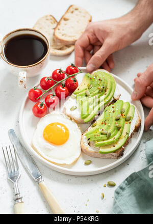 Mangiare sano colazione con avocado, uova, pane tostato e caffè. Mani tenendo la piastra con la prima colazione. Messa a fuoco selettiva, uno stile di vita sano concetto Foto Stock