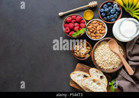 Sana colazione ingredienti nero su sfondo di calcestruzzo. Fiocchi di avena, di latte di mandorle, noci, frutta e bacche. Uno stile di vita sano, dieta e mangiare sano Foto Stock