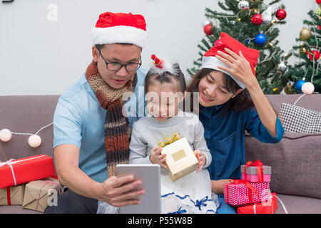 La famiglia felice Asia selfie con la compressa a divano,il padre e la madre usura santa claus hat dare il regalo di Natale scatola per bambino a casa xmas party,Holiday celeb Foto Stock