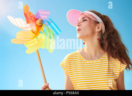 Montare sorridente donna in maglia gialla contro il cielo blu che soffia su girandola colorata Foto Stock