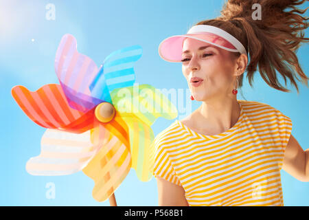 Felice fit donna in maglia gialla contro il cielo blu che soffia su girandola colorata Foto Stock