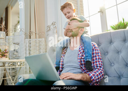 Ritratto di carino piccolo ragazzo che copre gli occhi del bel papà utilizzando computer portatile mentre è seduto sulle sue spalle e giocare in casa nella luce del sole Foto Stock
