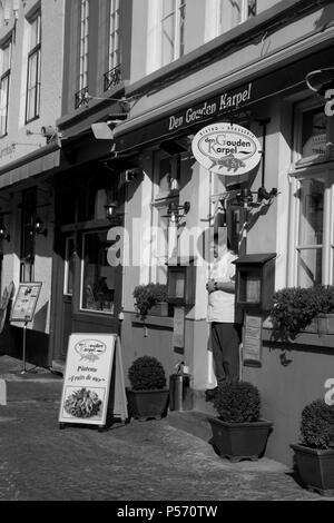 Huidenvettersplein, Brugge, Belgio: un cameriere sorge nella porta di Den Gouden Karpel ristorante. Versione in bianco e nero Foto Stock